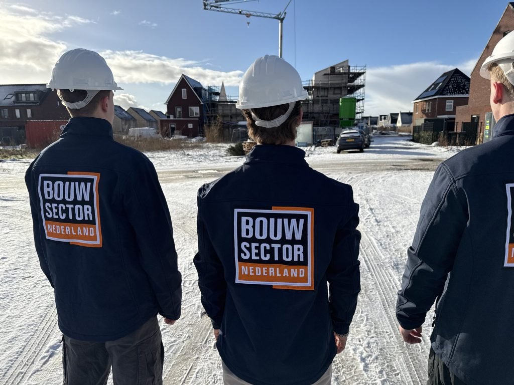 Drie mannen in werkjas met het logo van Bouwsector Nederland voor een rij huizen in aanbouw onder een blauwe lucht
